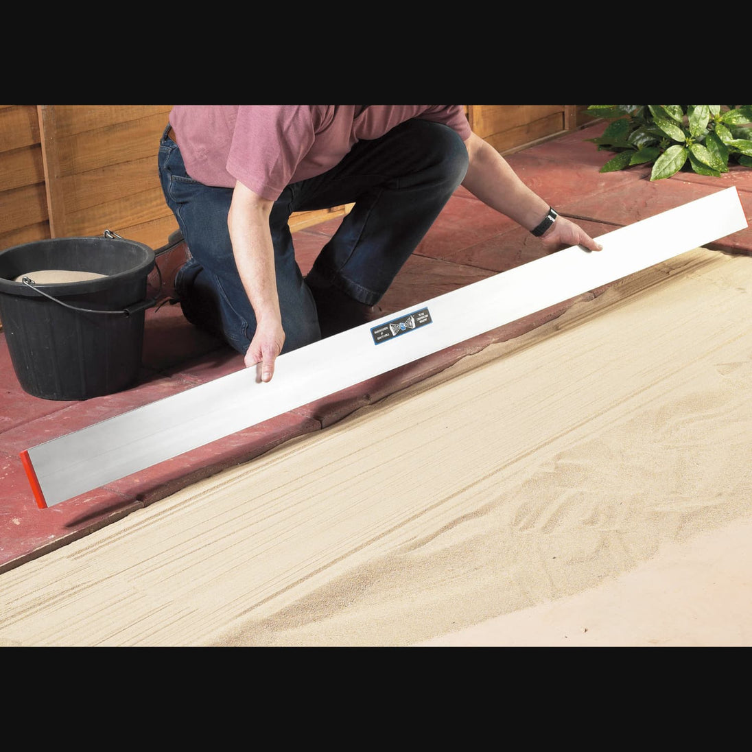 Man holding a long straight edge tool to level out sand