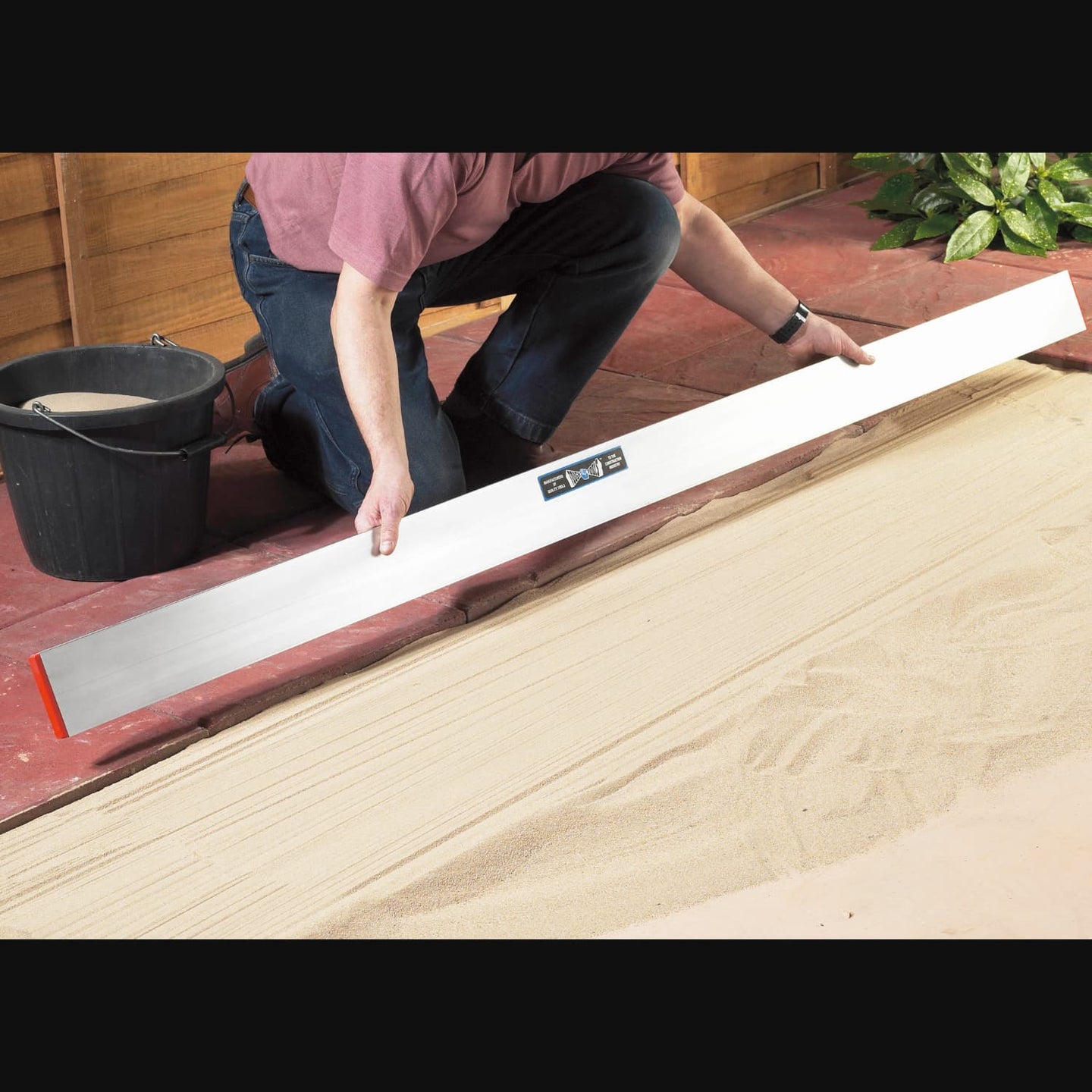 Man holding a long straight edge tool to level out sand